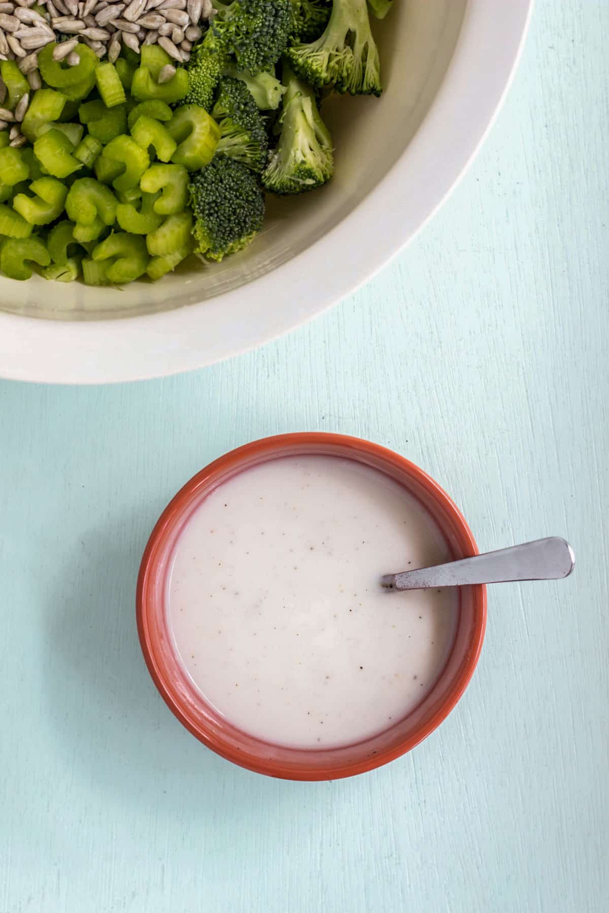 A small orange bowl of dressing with a spoon resting in it. A bowl with celery, broccoli and sunflower seeds is partially visible in the upper left corner.