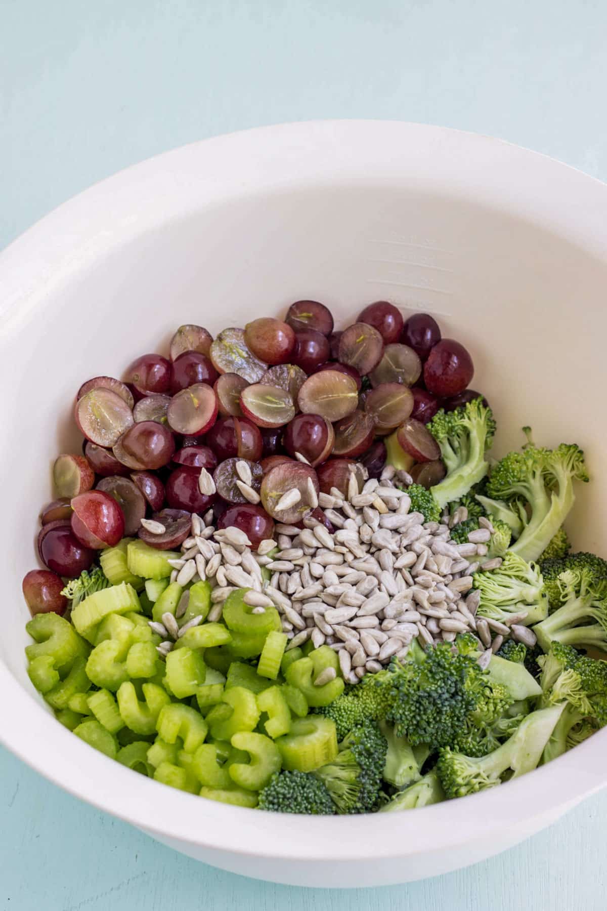 Fresh broccoli, chopped celery, grape halves and sunflower seeds in a large white bowl. The ingredients to make Vegan Broccoli Grape Salad.