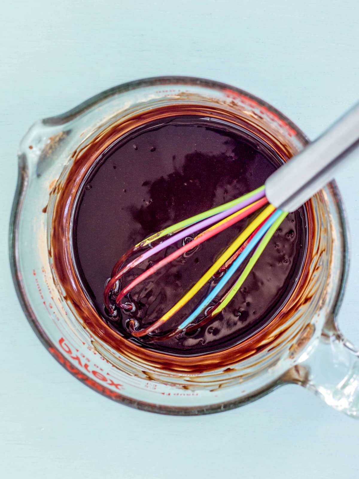 A large pyrex measuring cup of melted chocolate with a colorful whisk resting in it.