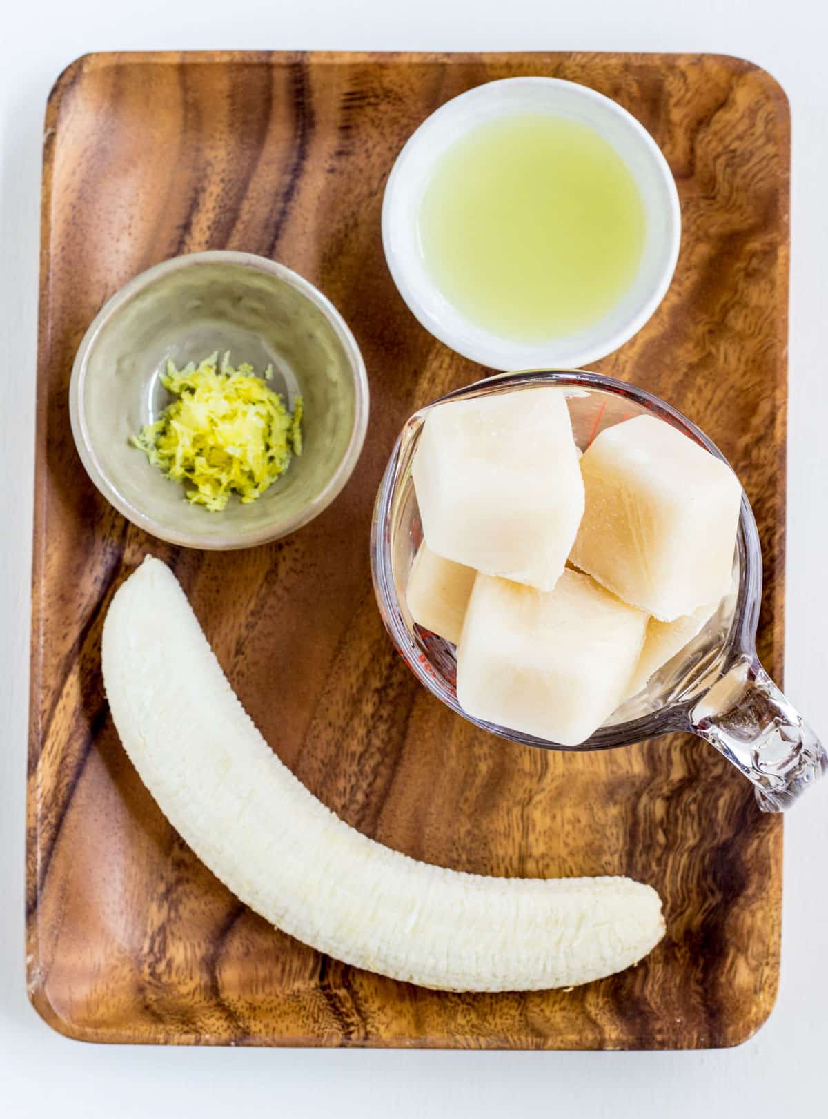 Lemon juice, zest, a whole frozen banana and non dairy milk ice cubes on a wooden plate.