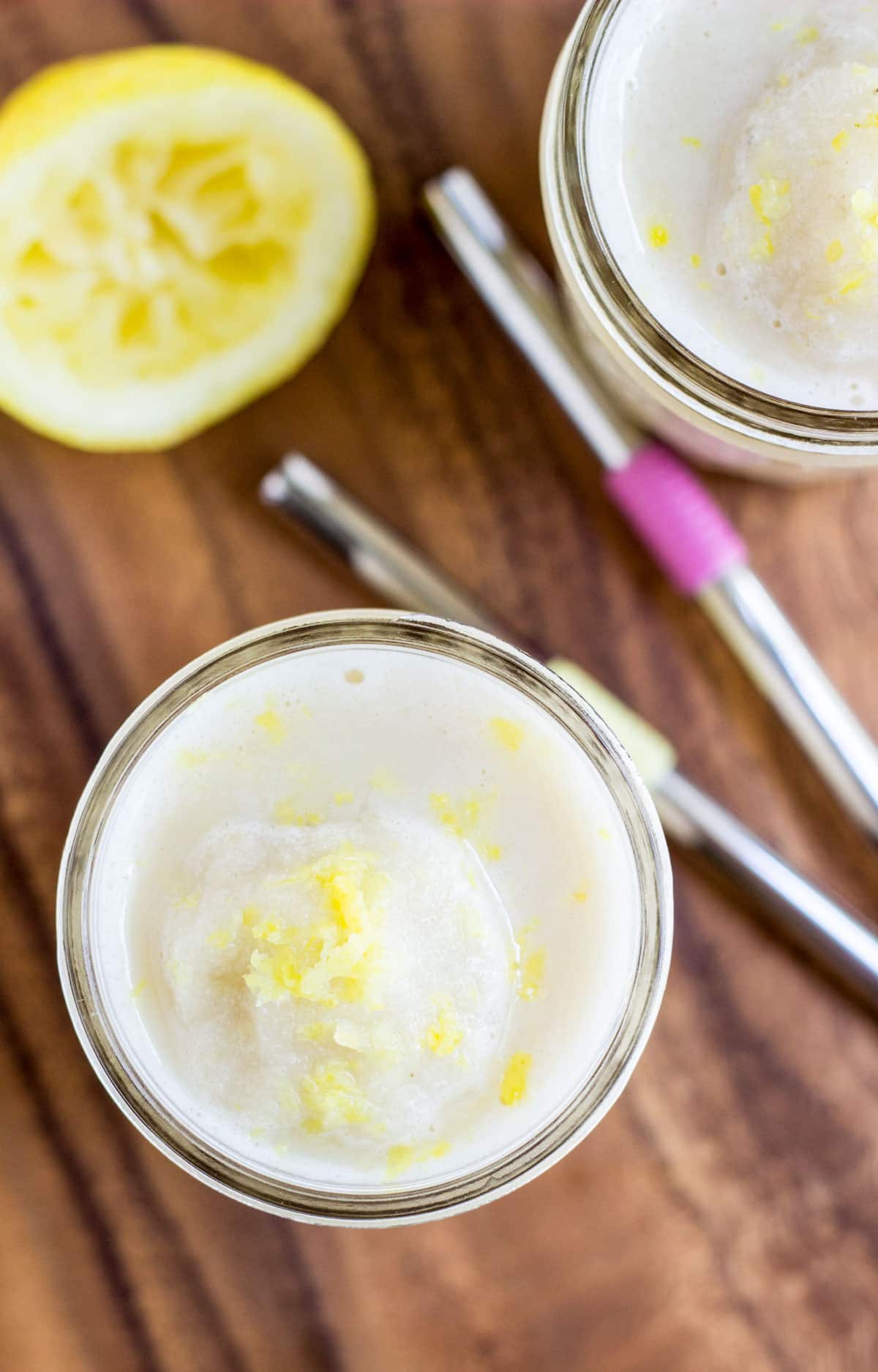 Top view of a glass of Creamy Vegan Lemonade Smoothie sitting on a wooden board. Two metal straws, a lemon half and another glass is partially visible.