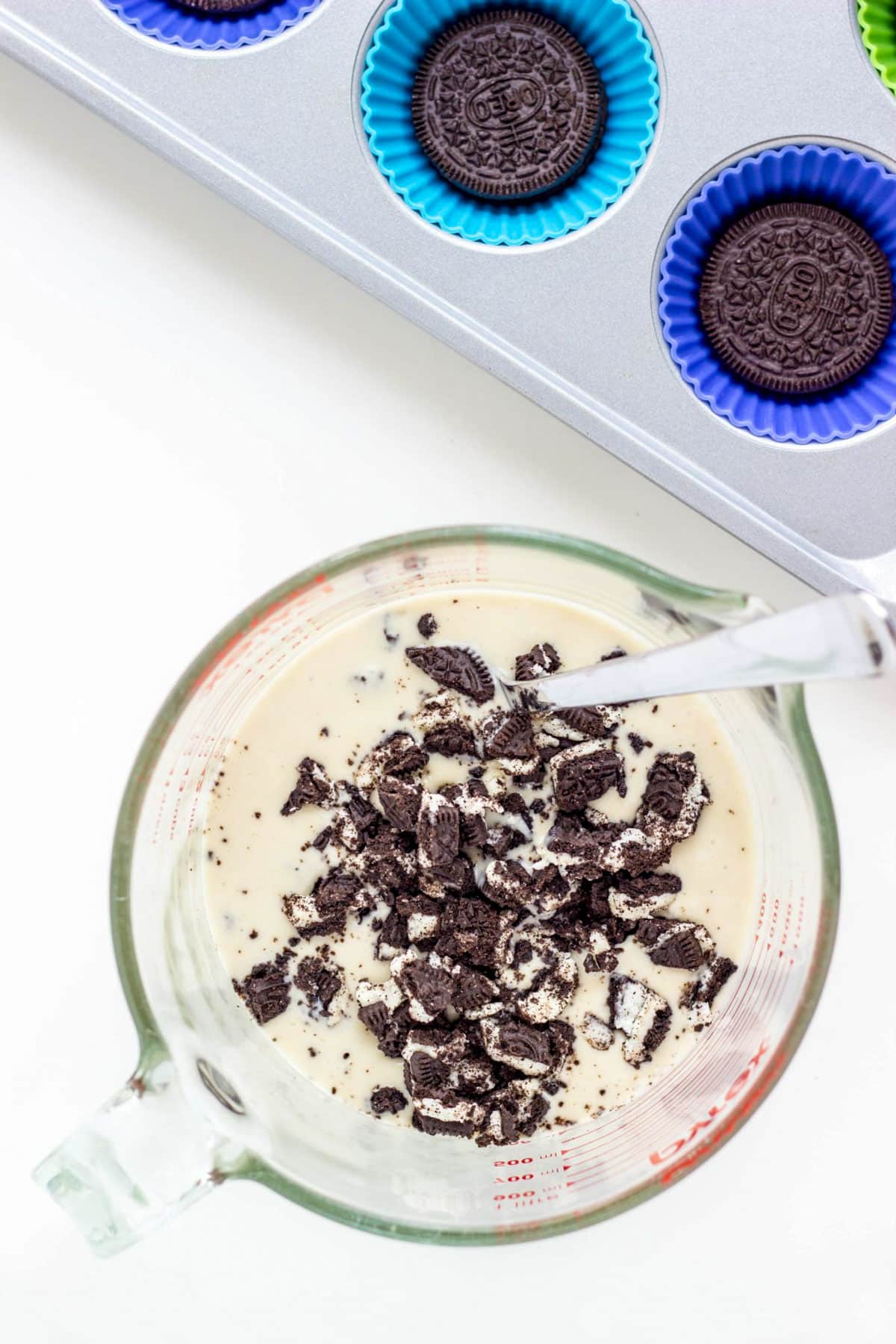 A measuring cup filled with ice cream base and sandwich cookies. A muffin pan with cookies in the wells is partially visible in the upper right of the frame.
