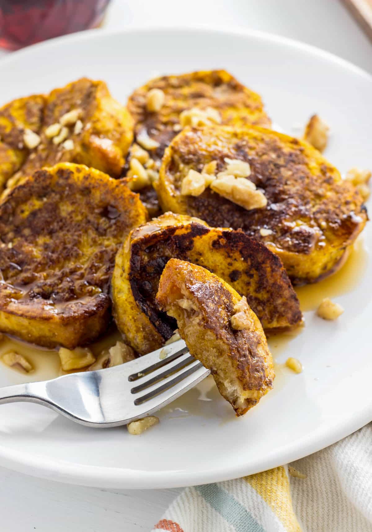Close up of a plate of Pumpkin Chia French Toast with chopped walnuts and maple syrup. There is a fork resting on the plate with a bite of French toast on it.