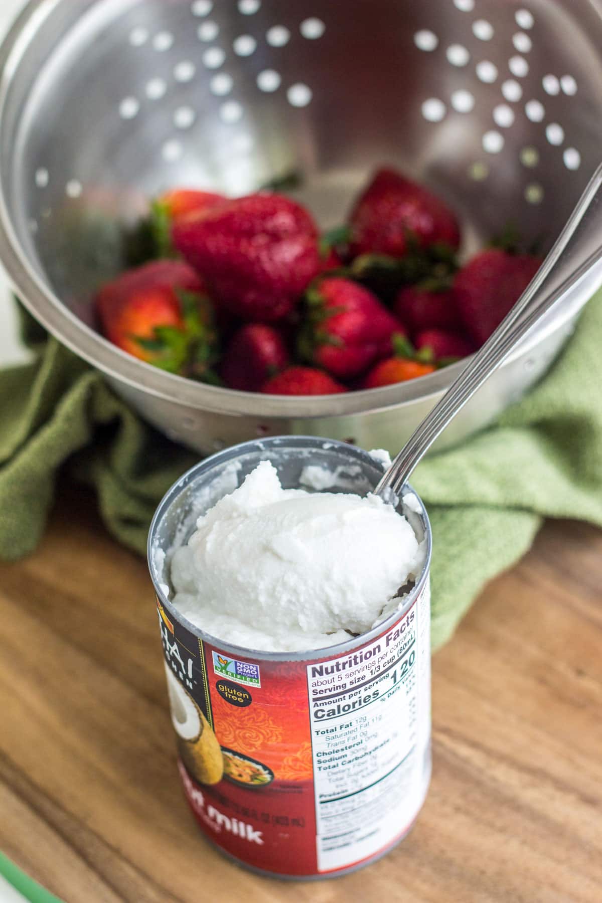 A can of coconut milk that has been refrigerated so the cream has separated and firmed up. There is a metal colander of strawberries visible in the background.
