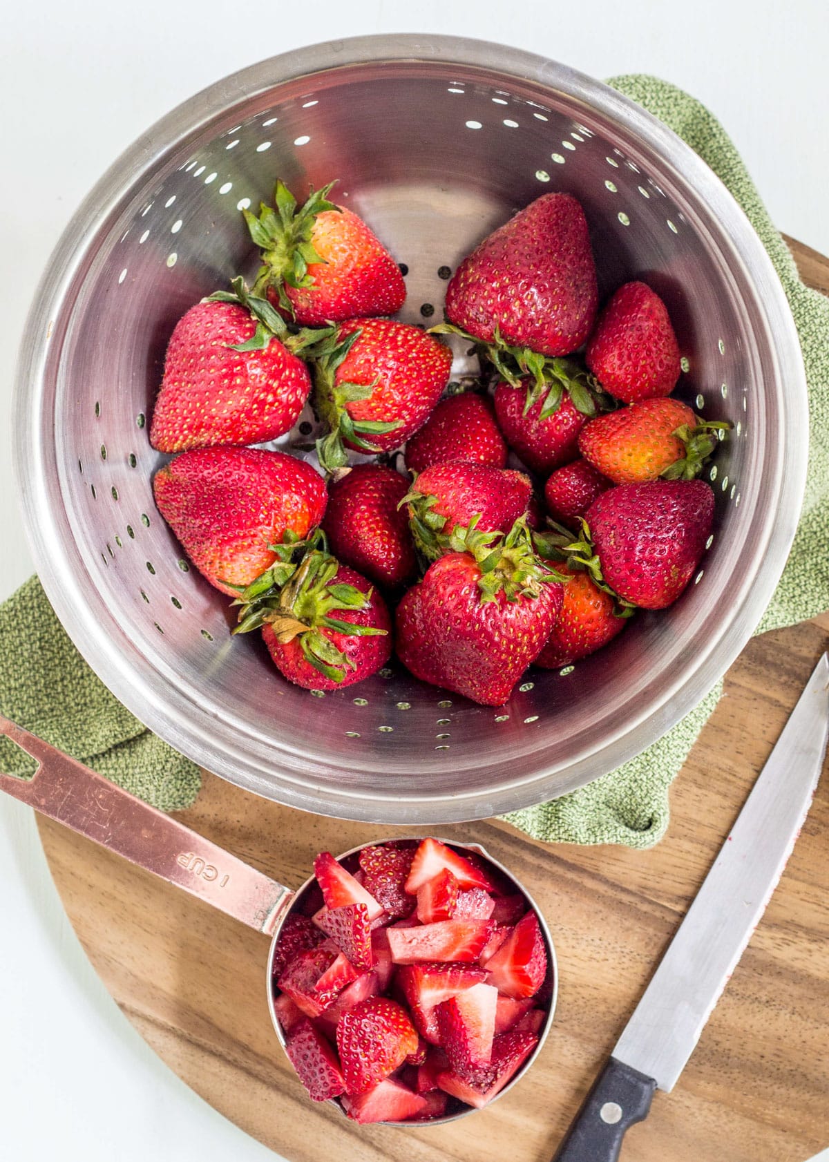 A measuring cup of chopped fresh strawberries sitting on a cutting board. There is also a metal colander of whole strawberries and a knife in the image.
