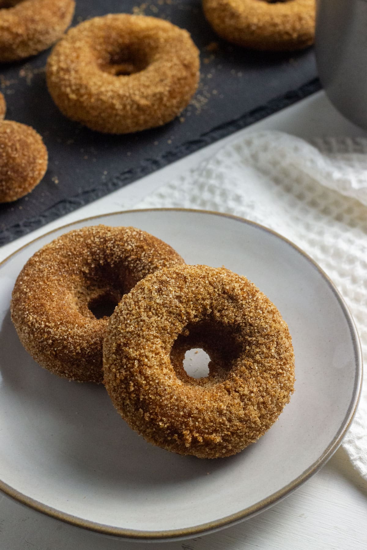 Two Vegan Apple Cider Doughnuts on a small plate. A slate tray with more doughnuts is partially visible in the background.