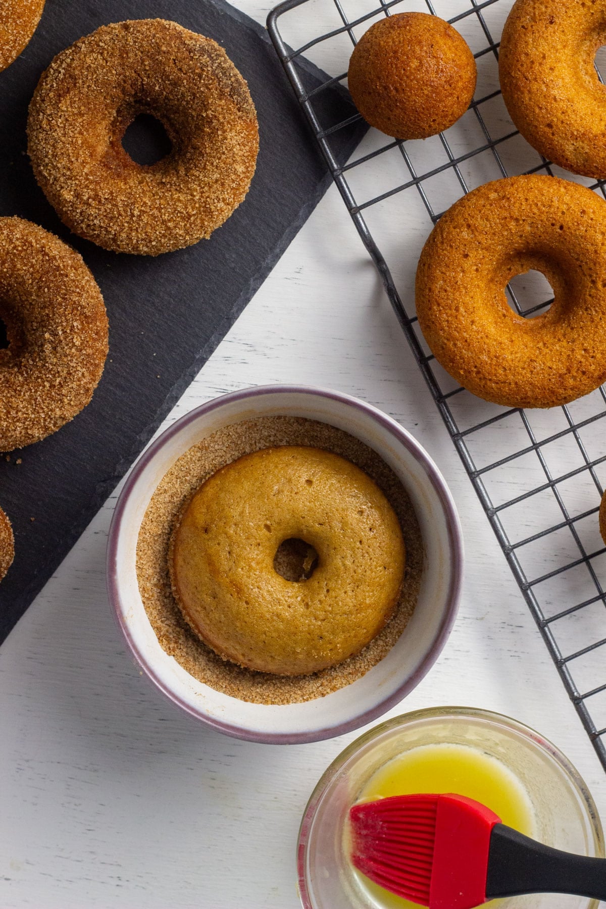 Overhead view of a doughnut resting in a small bowl of cinnamon sugar. There is a wire rack with uncoated doughnuts to the right and a slate tray with coated doughnuts to the left. In the bottom of the frame is a small bowl of melted non dairy butter and a rubber pastry brush.
