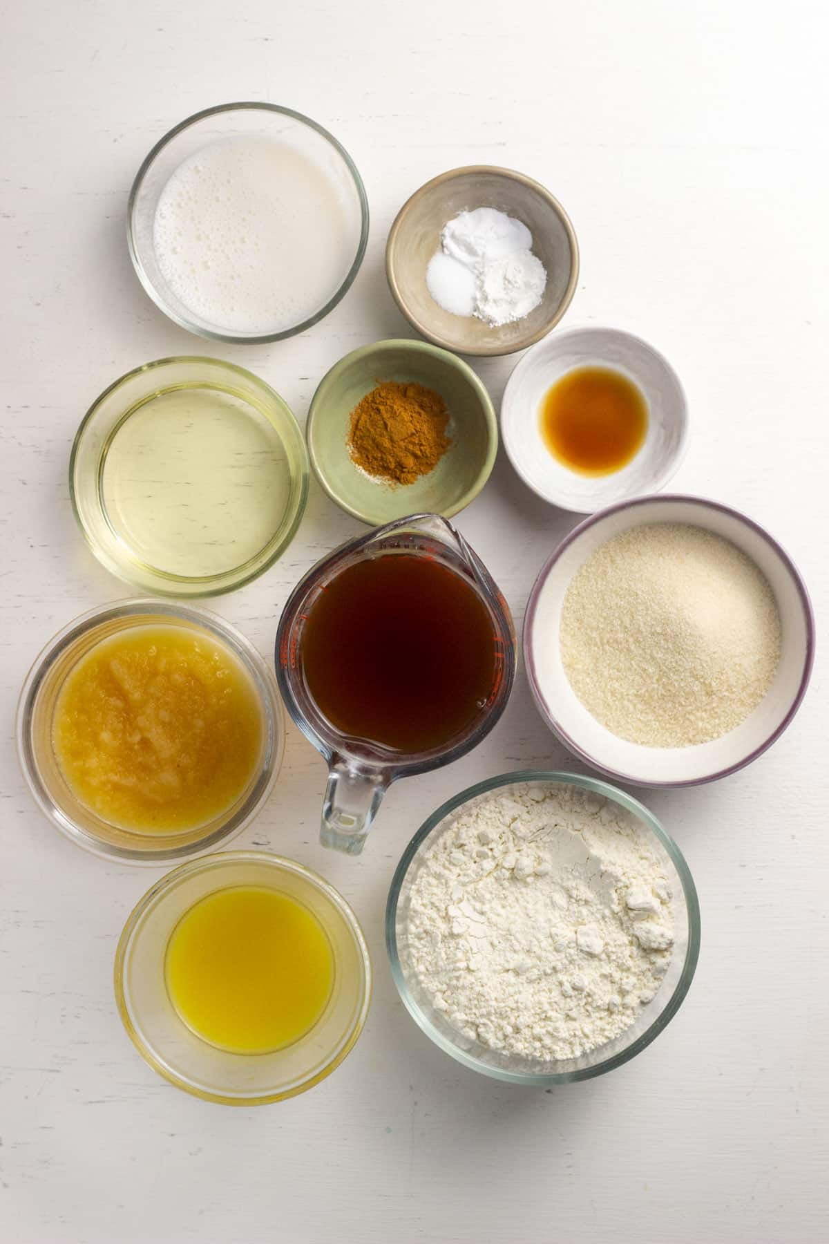 Ingredients to make Vegan Apple Cider Doughnuts. There are bowls of all purpose flour, sugar, vanilla, baking soda, baking powder, salt cinnamon, non dairy milk, oil, applesauce, melted nondairy butter and apple cider.