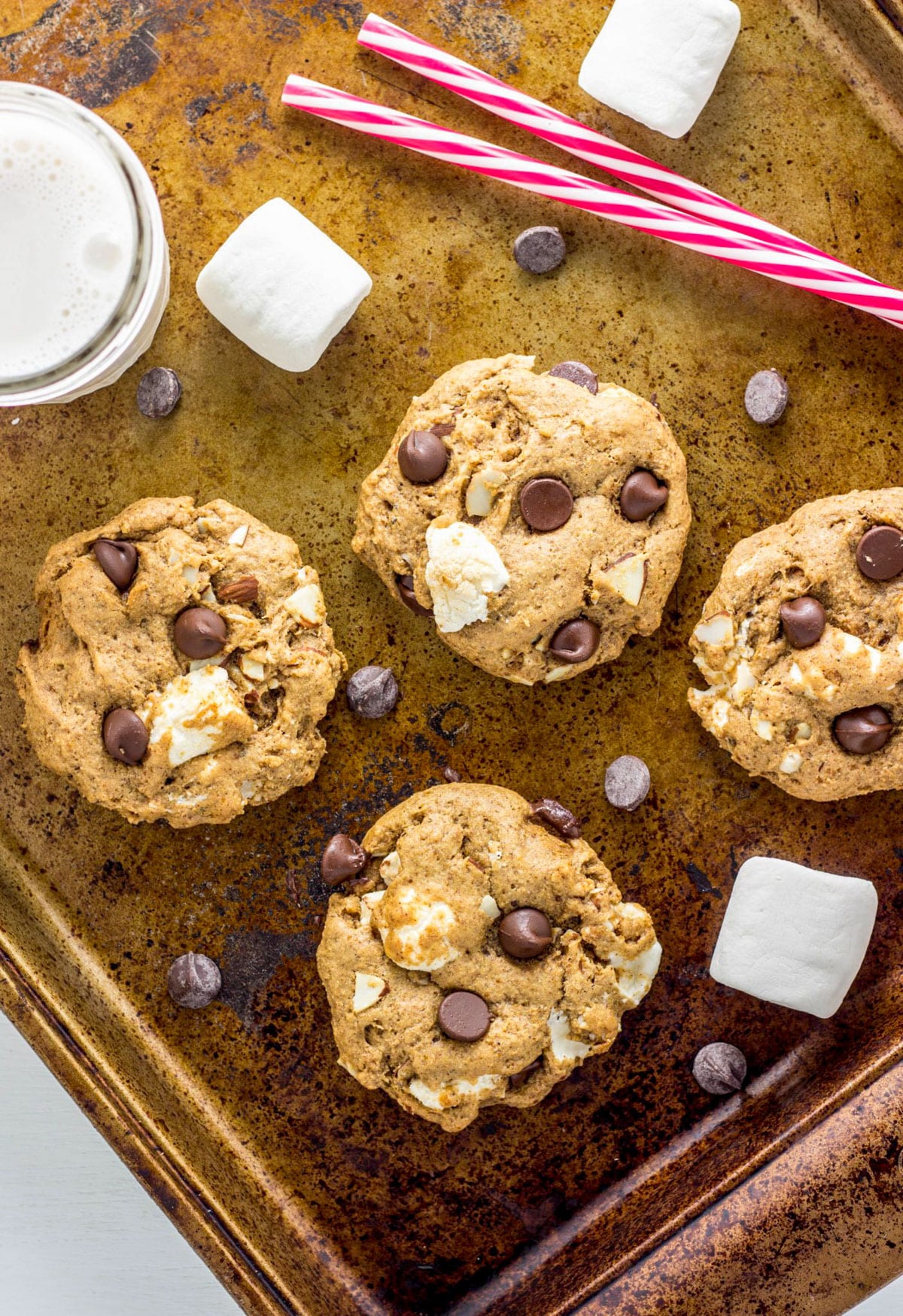 Vegan Rocky Road Cookies on a baking sheet with marshmallows and chocolate chips scattered around. A glass of almond milk is also partially in view.