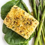 Top view of Baked Lemon & Herb Crusted Tofu on a bed of spinach and a side of asparagus on a white plate