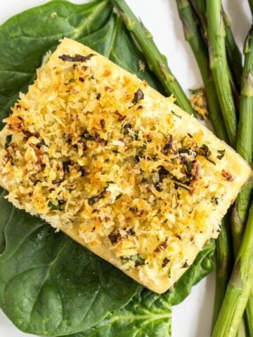 Top view of Baked Lemon & Herb Crusted Tofu on a bed of spinach and a side of asparagus on a white plate