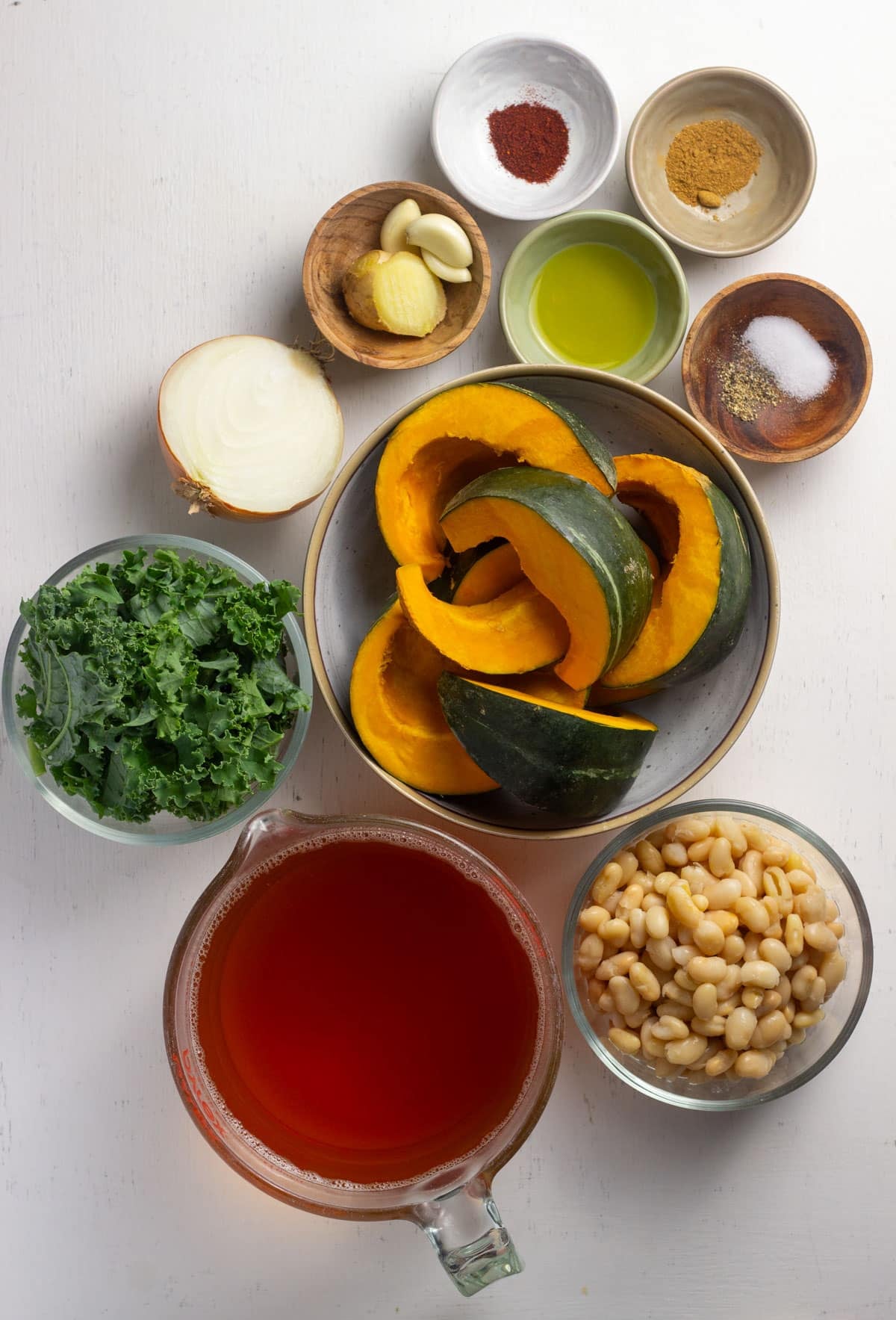 Ingredients to make Kabocha White Bean Soup. There are bowls of vegetable broth, white beans, kabocha squash wedges, kale, salt, pepper. cumin, smoked paprika, olive oil, fresh ginger and garlic cloves.