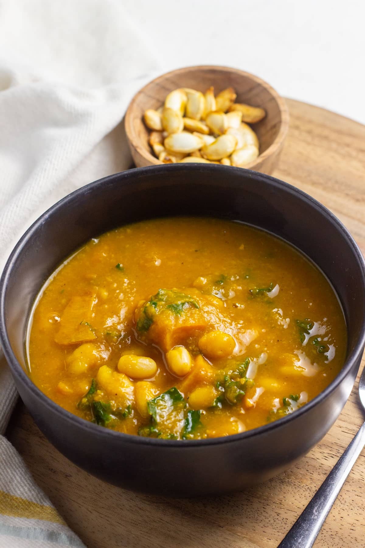 A black bowl of Kabocha White Bean Soup. In the background there is a small wooden bowl of roasted seeds from the kabocha squash.