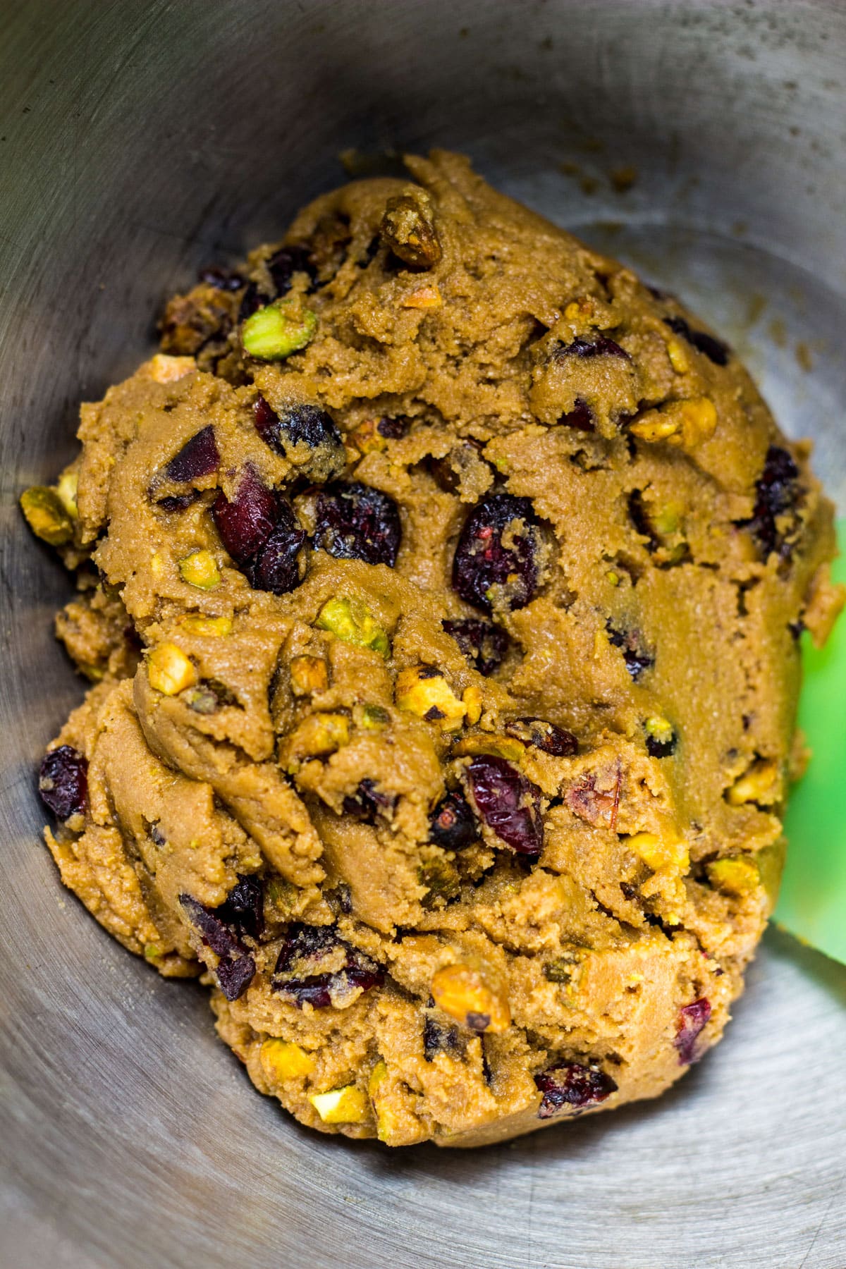 Close up of dough to make Vegan Cranberry Pistachio Cookies in a metal mixing bowl with a spatula to the side.