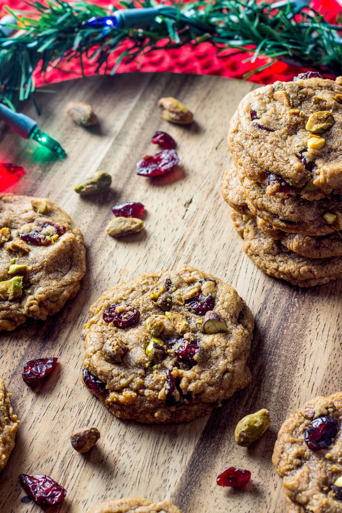 Vegan Cranberry Pistachio Cookies on a wooden board. There are dried cranberries and shelled pistachios around the cookies and Christmas lights are in the top of the frame.