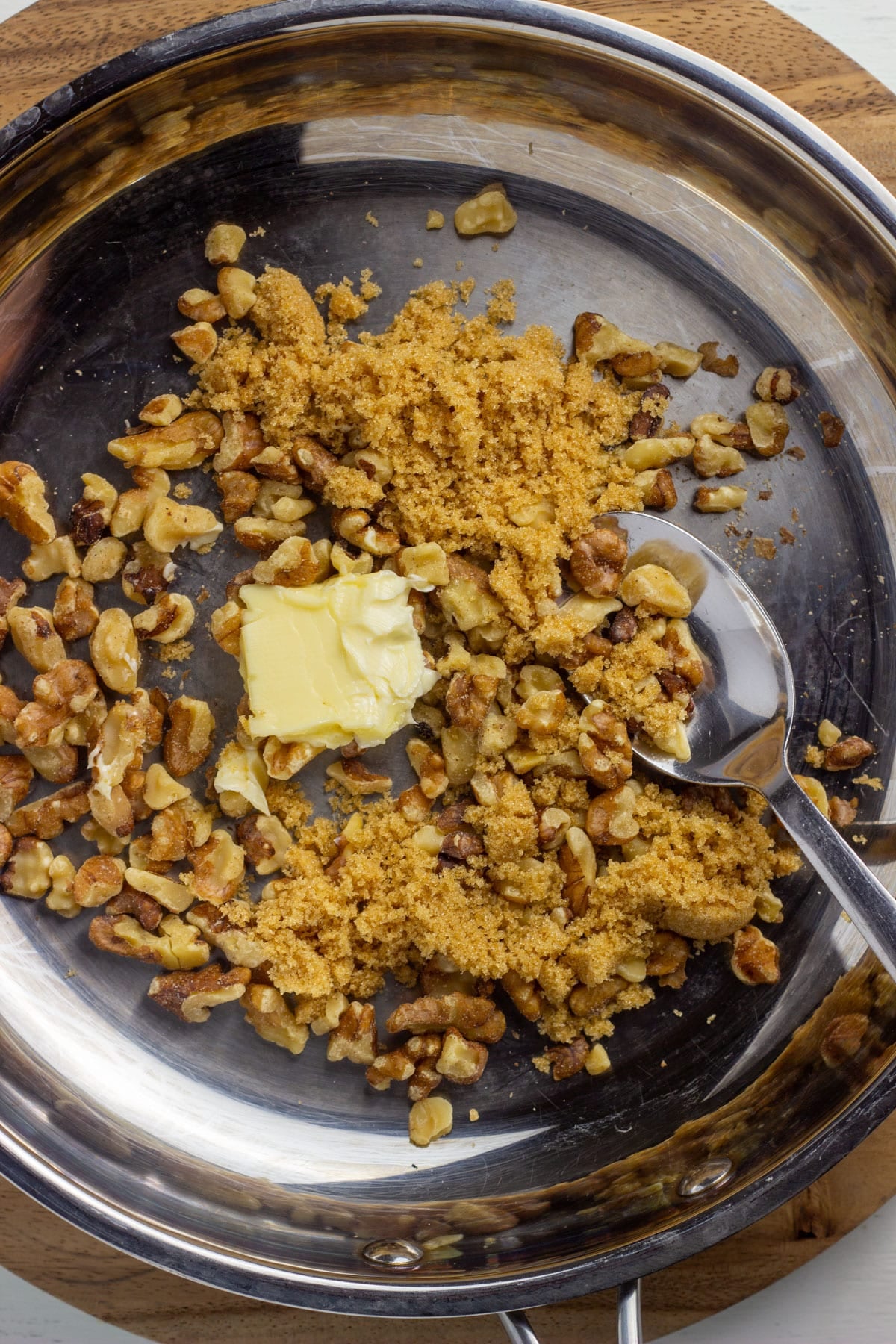 Walnuts, brown sugar and vegan butter in a skillet. A spoon is on the right side resting in the pan.